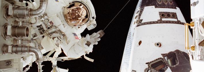 An astronaut in a white spacesuit appears on the left side of the image near a part. On the right in the back is part of the space shuttle Endeavour.