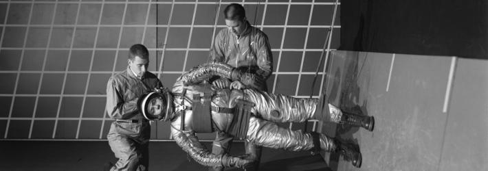 In this black and white photo, two people adjust a 1960s-era spacesuit on a person who is suspended by wires. This third person is parallel to the floor, with his feet on the wall on the right.