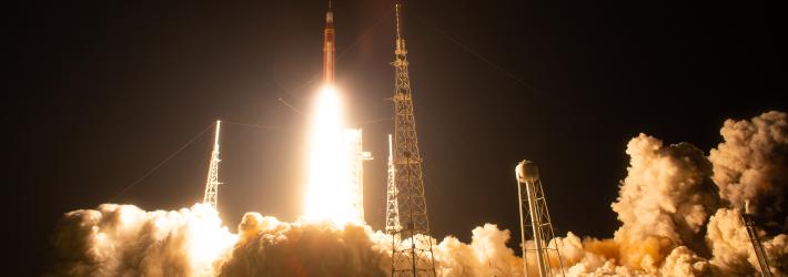 An orange rocket lifts off from a launchpad. The sky is black, but the fire coming from the engines lights up the surrounding area, highlighting clouds of white vapor spreading across the ground.