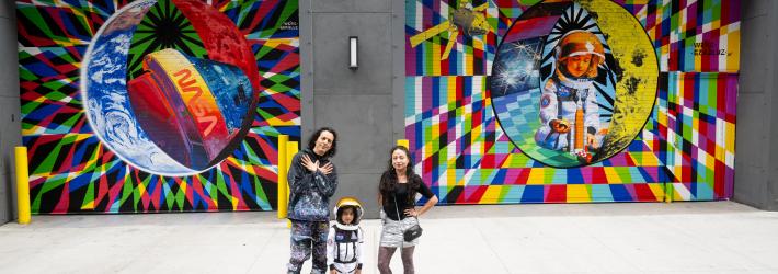 Two adults and a child pose for a photo in front of a colorful space-themed mural. The child wears an astronaut costume.