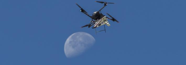 A white, blue, and red probe attached to a rotor with four blades flies in the blue sky, just above the Moon.