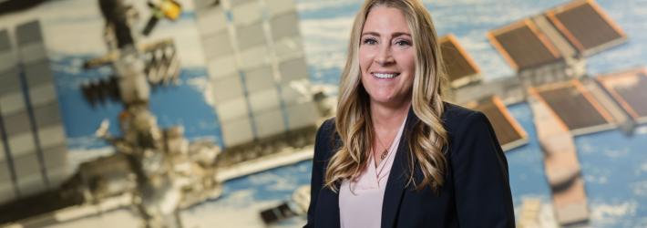 Dana Weigel stands and smiles in front of a large wall mural of the International Space Station. She has wavy blond hair and is wearing a dark blue jacket over a pink blouse with a circular necklace around her neck.