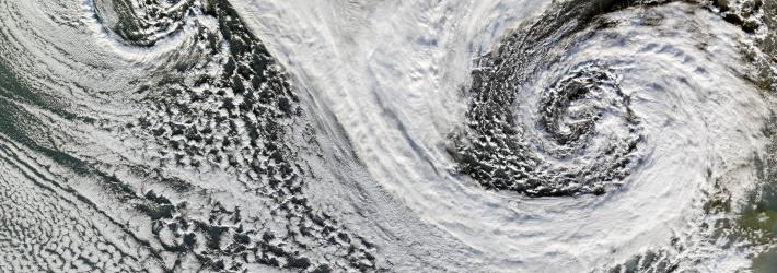 Two cyclones swirl south of Iceland in this satellite image. While the two storms' distinctive spiral patterns are clearly visible at left and right, white clouds cover most of the image. Some of Iceland can be seen at the top middle of the image, as well as part of Scotland at bottom right.