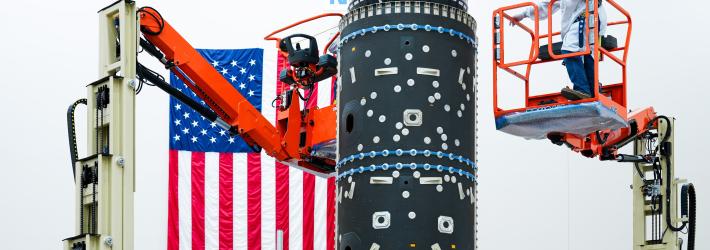 A black piece of equipment with small silver pieces all over it is at the center of this image. This is what houses Gateway's fuel tanks. A person in a blue hairnet, mask, white lab coat, and jeans stands on an orange lift that is several feet in the air. Another person in similar clothing is at the bottom of the piece of equipment at the center of this image. An American flag hangs vertically on the wall.