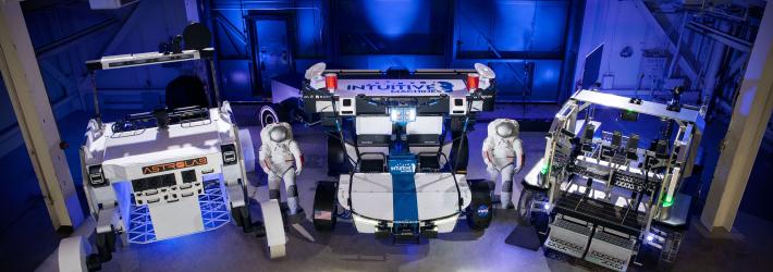 Three lunar terrain vehicles side by side inside of a large chamber with dramatic lighting. Astrolab's lunar terrain vehicle is white with the their orange logo on the front of the rover. Intuitive Machines' lunar terrain vehicle is white with a blue stripe on the front and their logo on the top of their rover. Lunar Outpost's lunar terrain vehicle is grey with green lights.