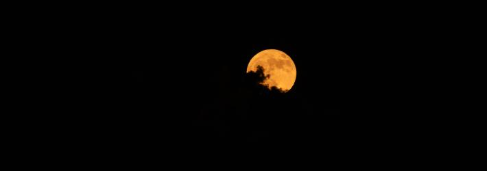 An orange full moon is the only thing you can see in this image. The sky is black, and the outline of some clouds are visible against the Moon.