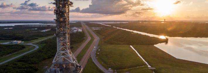 A large rectangular structure - the mobile launcher - rests atop a moving rectangular base - the crawler-transporter. In this aerial shot, they are moving along a road. Green grass covers the surrounding area.