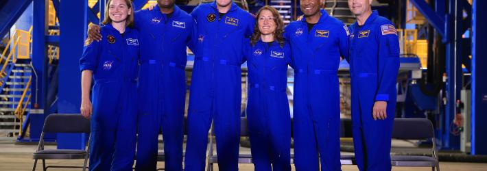 Six astronauts, all in blue jumpsuits, pose together for a photo.