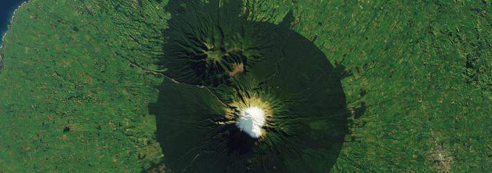 An overhead view of Mount Taranaki from a satellite. A white peak (a snow-capped mountain) is circled by dark green forest. The rest of the land around it is a bright green.