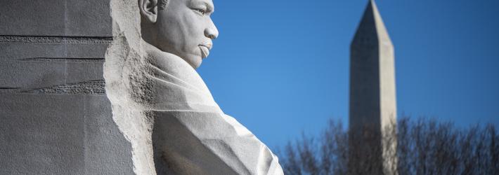 A close-up image of a light gray stone statue of Martin Luther King, Jr. in Washington. The Washington Monument is visible in the background, behind the leafless branches of a tree.