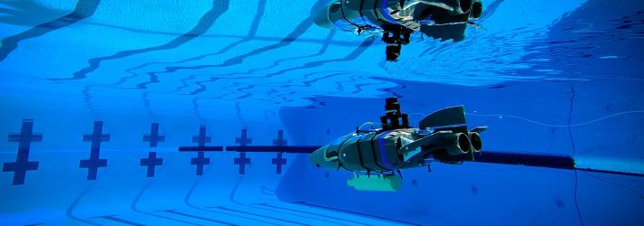 A small gray plastic robot shaped like a wedge (with propellers and flaps for steering) glides through the blue waters of a competition swimming pool.